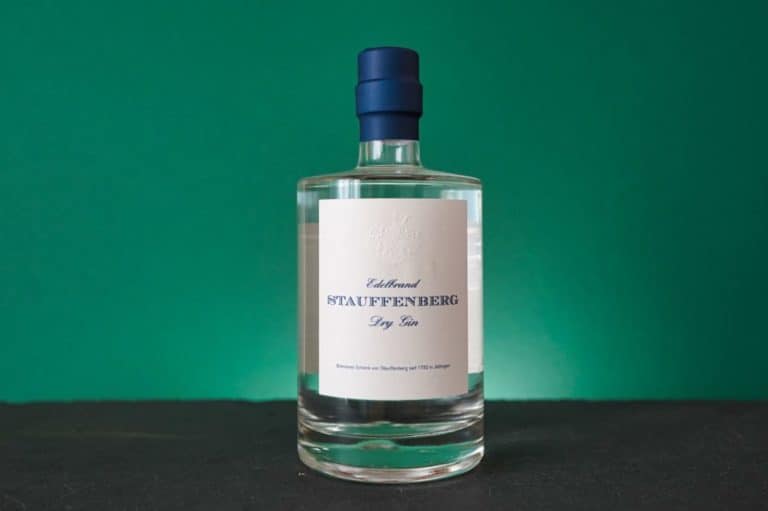 Eine klare Glasflasche Stauffenberg Gin mit blauem Verschluss steht auf einer dunklen Oberfläche vor einem grünen Hintergrund. Das weiße Etikett ist mit blauer Schrift bedruckt.