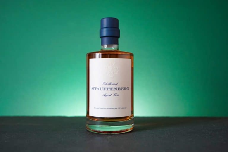 Eine Flasche „Gin Stauffenberg Dry Gin“ mit weißem Etikett und blauem Verschluss steht auf einer dunklen Oberfläche vor einem grünen Hintergrund.