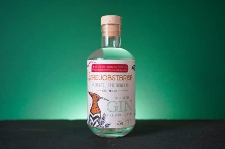 Eine Flasche „Streuobstbrise Gin“ aus Freiburg mit einer Vogelillustration auf dem Etikett steht auf einer dunklen Fläche vor einem grünen Hintergrund.