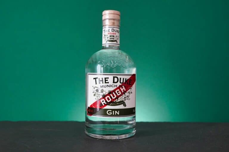 Auf einer dunklen Fläche mit grünem Hintergrund steht eine klare Glasflasche mit der Aufschrift „The Duke Rough Gin“. Das Etikett zeigt florale Motive und einen roten Streifen mit der Aufschrift „Rough“, der die unverwechselbare Essenz des „The Duke Munich Rough Gin“ einfängt.
