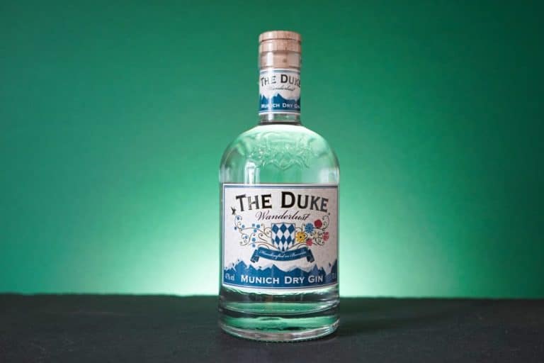 Eine durchsichtige Flasche „The Duke Munich Dry Gin“ steht neben einer Flasche „The Duke Wanderlust Gin“ mit blau-weißem Etikett vor einem grünen Farbverlauf auf einem dunklen Untergrund.