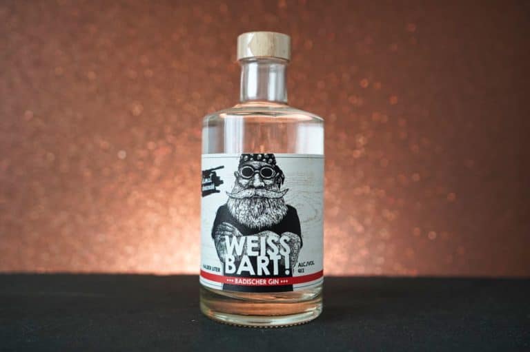 Eine klare Glasflasche mit „Weissbart Gin“, auch bekannt als „Weiss Bart Badischer Gin“, ist mit einem Etikett versehen, auf dem ein bärtiger Mann mit Schutzbrille vor einem glitzernden, kupferfarbenen Hintergrund abgebildet ist.