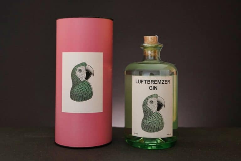 Neben einer Flasche „Luftbremzer Gin“ mit einer Papageienabbildung auf dem Etikett steht eine dazu passende rosa zylindrische Schachtel; beide sind mit dem unverwechselbaren „Luftbremzer Gin“-Logo versehen, das sich vor einem schlichten schwarzen Hintergrund abhebt.