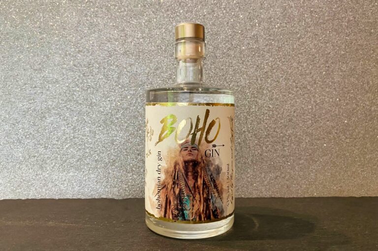 Eine Flasche Boho Gin (Bohemian Dry Gin) mit einem Etikett, auf dem eine Person mit langen Haaren und Schmuckstücken vor einem funkelnden, strukturierten Hintergrund abgebildet ist.