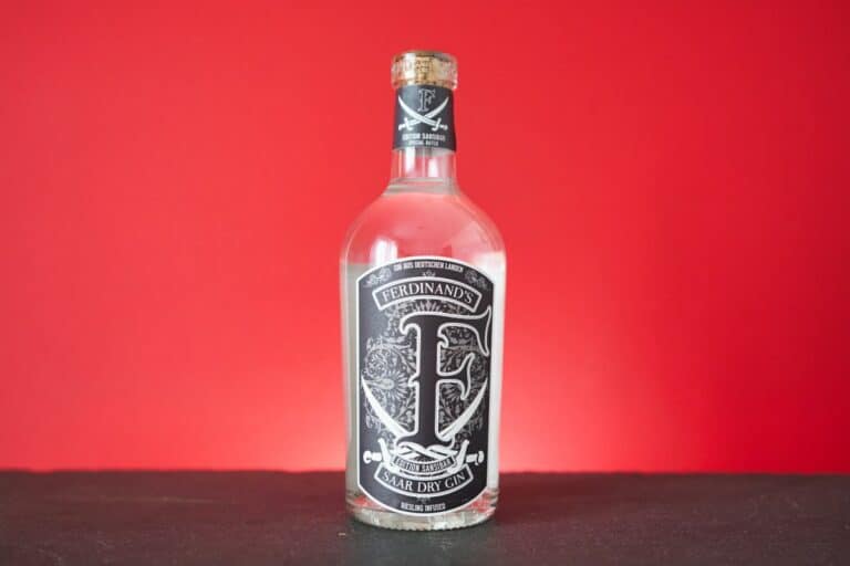 Eine klare Glasflasche „Ferdinands Saar Dry Gin“ mit einem schwarz-weißen Etikett steht auf einer schwarzen Fläche vor einem leuchtend roten Hintergrund und unterstreicht die unverwechselbare Eleganz von Ferdinands Gin.