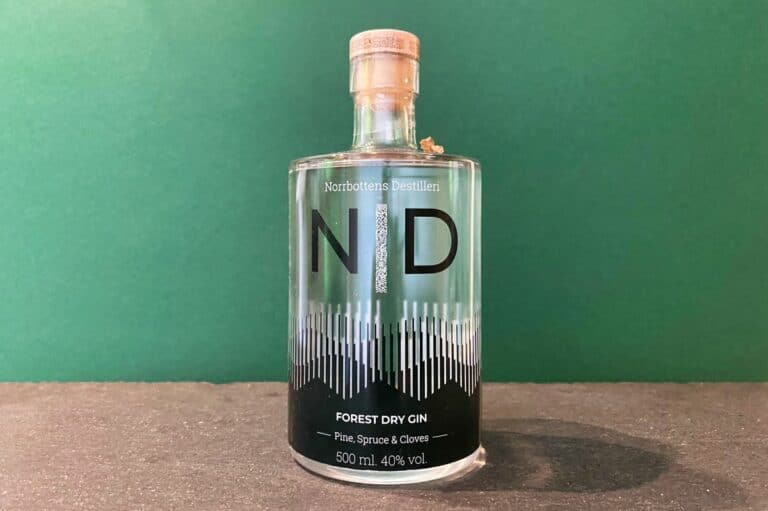 Eine durchsichtige Flasche „Norrbottens Destilleri Forest Dry Gin“ mit Korkverschluss steht auf einer dunklen Oberfläche vor einem grünen Hintergrund. Das Etikett hebt diesen außergewöhnlichen Forest Gin hervor, erwähnt Aromen von Kiefer, Fichte und Nelken und gibt 500 ml sowie 40 % Vol. an.