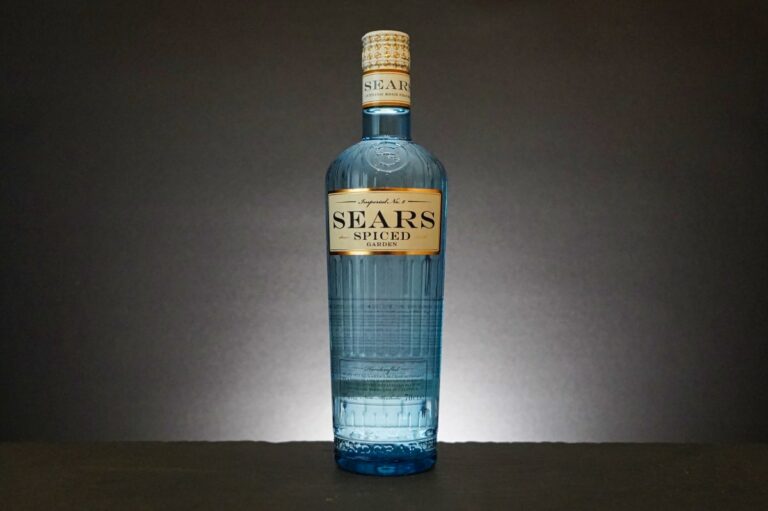 Eine blaue Glasflasche mit der Aufschrift „SEARS SPICED“ steht aufrecht vor einem dunklen Hintergrund, hinter ihr ist ein heller Lichtfleck zu sehen, der den raffinierten Charakter von Gin Sears verkörpert.