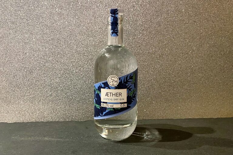 Eine durchsichtige Flasche „Aether Leipzig Dry Gin“ mit einem blau-weißen Etikett steht auf einer dunklen Fläche vor einem funkelnden silbernen Hintergrund.