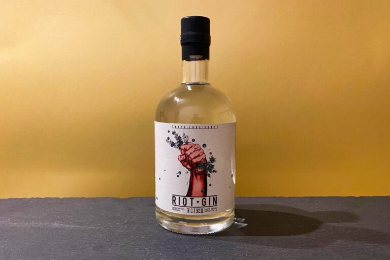Eine durchsichtige Flasche „Riot Gin“ mit einem Etikett, auf dem stolz eine erhobene Faust zu sehen ist, die Kräuter umklammert, steht vor einem schlichten gelben Hintergrund auf einer dunklen Fläche.