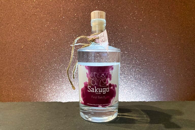 Eine durchsichtige Flasche mit der Aufschrift „Sakugo First Batch“, einer violetten Grafik, einem Korkverschluss und einem Etikett aus Schnur steht auf einer dunklen Fläche vor einem glitzernden braunen Hintergrund und bringt den raffinierten Charakter des Sakugo Dry Gin elegant zur Geltung.