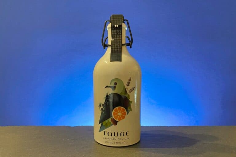Eine weiße Flasche „Taube Bavarian Dry Gin“, bekannt als „Taube Gin“, mit einer Illustration eines Vogels und einer Zitrusfrucht auf dem Etikett vor blauem Hintergrund.