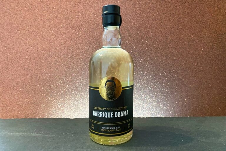 Eine durchsichtige Gin-Flasche mit der Aufschrift „Barrique Obama Single Cask Gin“ und einem schwarz-goldenen Design mit einem Porträt steht auf einer dunklen Fläche vor einem funkelnden braunen Hintergrund.