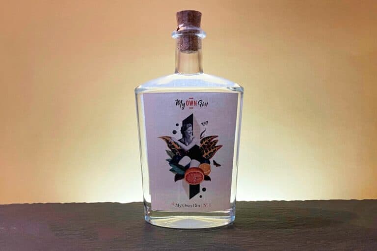 Eine klare Glasflasche mit Korkverschluss, die mit „My Own Gin No. 1“ beschriftet ist und auf der ein abstraktes Motiv mit Früchten und Pflanzen zu sehen ist, steht vor einem warmen beigen Hintergrund auf einer dunklen Fläche.
