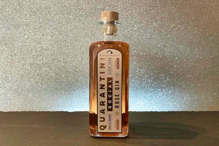 Eine Flasche „Quarantini Gin Social Rose“ steht auf einer dunklen Fläche vor einem glitzernden, silbernen Hintergrund. Die Flasche hat einen Holzverschluss und ein stilvolles, modernes Etikett.