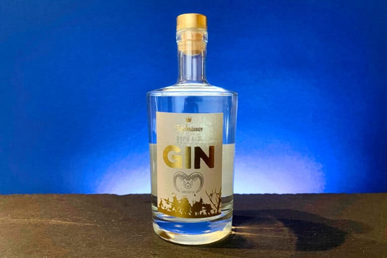 Eine durchsichtige Gin-Flasche mit einem goldenen Etikett, auf dem „GIN“ steht, steht auf einer dunklen Fläche vor blauem Hintergrund und wird von hinten beleuchtet – so wird der kühne Charakter von „Tafelmänner – Bock auf Gin“ eingefangen.