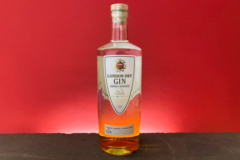 Eine Flasche Rewe Gin – ein klassischer London Dry mit goldfarbener Flüssigkeit – steht vor einem leuchtend roten Hintergrund auf einer dunklen Unterlage. Das Etikett ist weiß und mit goldenen und roten Akzenten versehen.