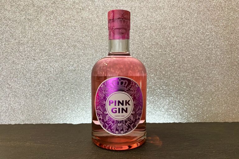 Eine durchsichtige Glasflasche mit rosa Gin und einem violett-silbernen Etikett steht auf einer dunklen Fläche vor einem funkelnden, strukturierten hellgrauen Hintergrund – perfekt für alle, die die Premium-Gin-Auswahl von Rewe entdecken möchten.