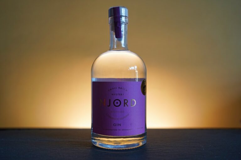 Eine durchsichtige Flasche „NJORD Distilled Comeback Quinine Gin“ mit einem violetten Etikett steht auf einer dunklen Oberfläche vor einem gelb-orangefarbenen Hintergrund mit Farbverlauf und wird von hinten beleuchtet.