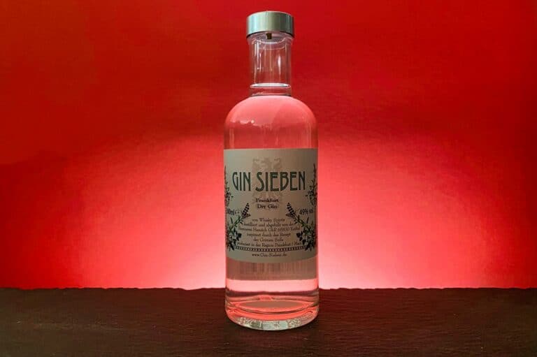 Auf einer dunklen Fläche vor rotem Hintergrund steht eine durchsichtige Flasche mit der Aufschrift „Gin Sieben“, die von hinten beleuchtet wird, um die Transparenz des darin enthaltenen Gin Sieben hervorzuheben.