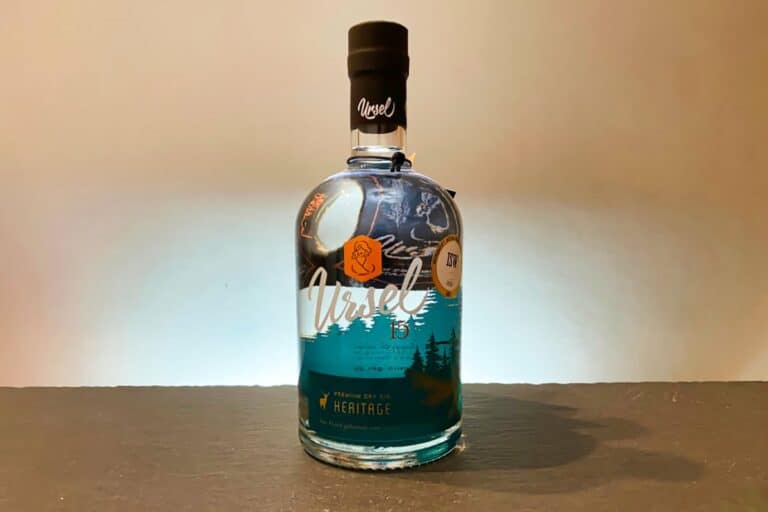 Eine Flasche Ursel Gin Heritage steht auf einer dunklen Unterlage vor einem hellen Hintergrund. Das Etikett zieren das ikonische Fuchs-Logo und Waldmotive, während die Flüssigkeit im Inneren eine charakteristische blaue Färbung aufweist.