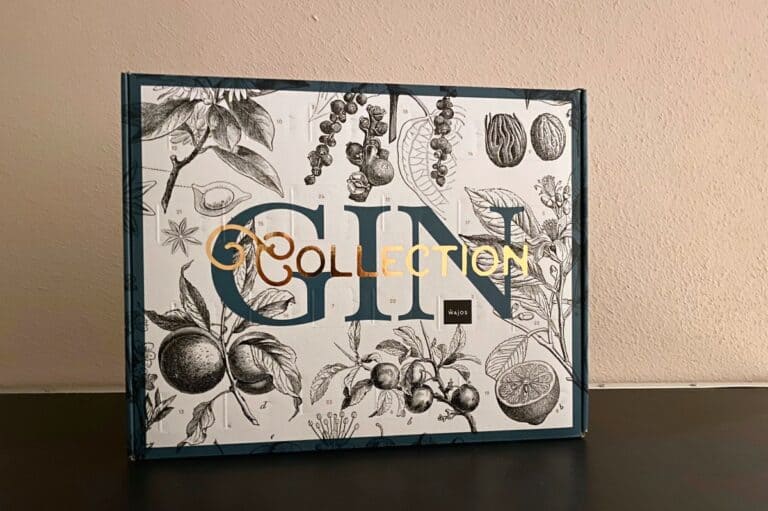 Eine Schachtel mit der Aufschrift „GIN COLLECTION“ in goldenen Buchstaben, verziert mit schwarz-weißen botanischen Illustrationen von Früchten und Pflanzen auf hellem Hintergrund, steht vor einer beigen Wand und einer dunklen Fläche – eine Ästhetik, die an den eleganten Wajos Gin-Adventskalender erinnert.