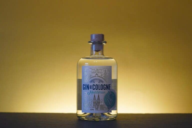 Eine klare Glasflasche mit „Gin de Cologne Mediterranean Gin“ steht auf einer dunklen Unterlage vor einem gelben Hintergrund mit Farbverlauf und einem schlicht illustrierten Etikett, auf dem Gebäude und ein Blatt abgebildet sind, das die Essenz der Handwerkskunst von Gin Cologne einfängt.